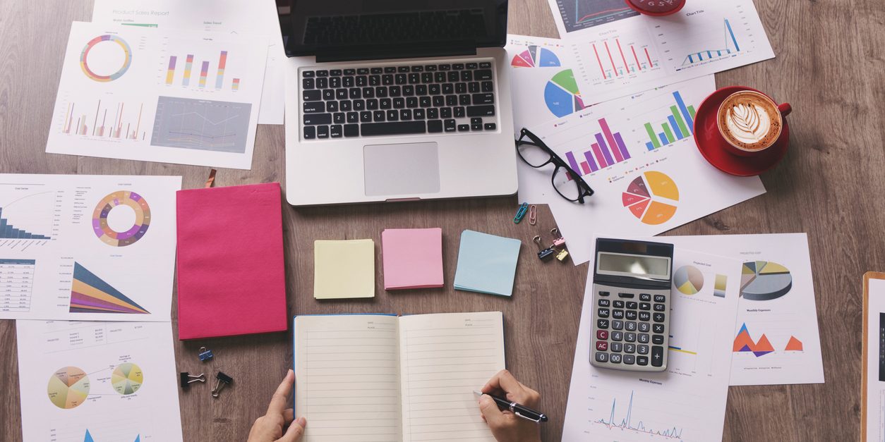 Top view of woman's hand writing on a notebook with laptop, calculator, a cup of coffee , financial chart and other stationary.